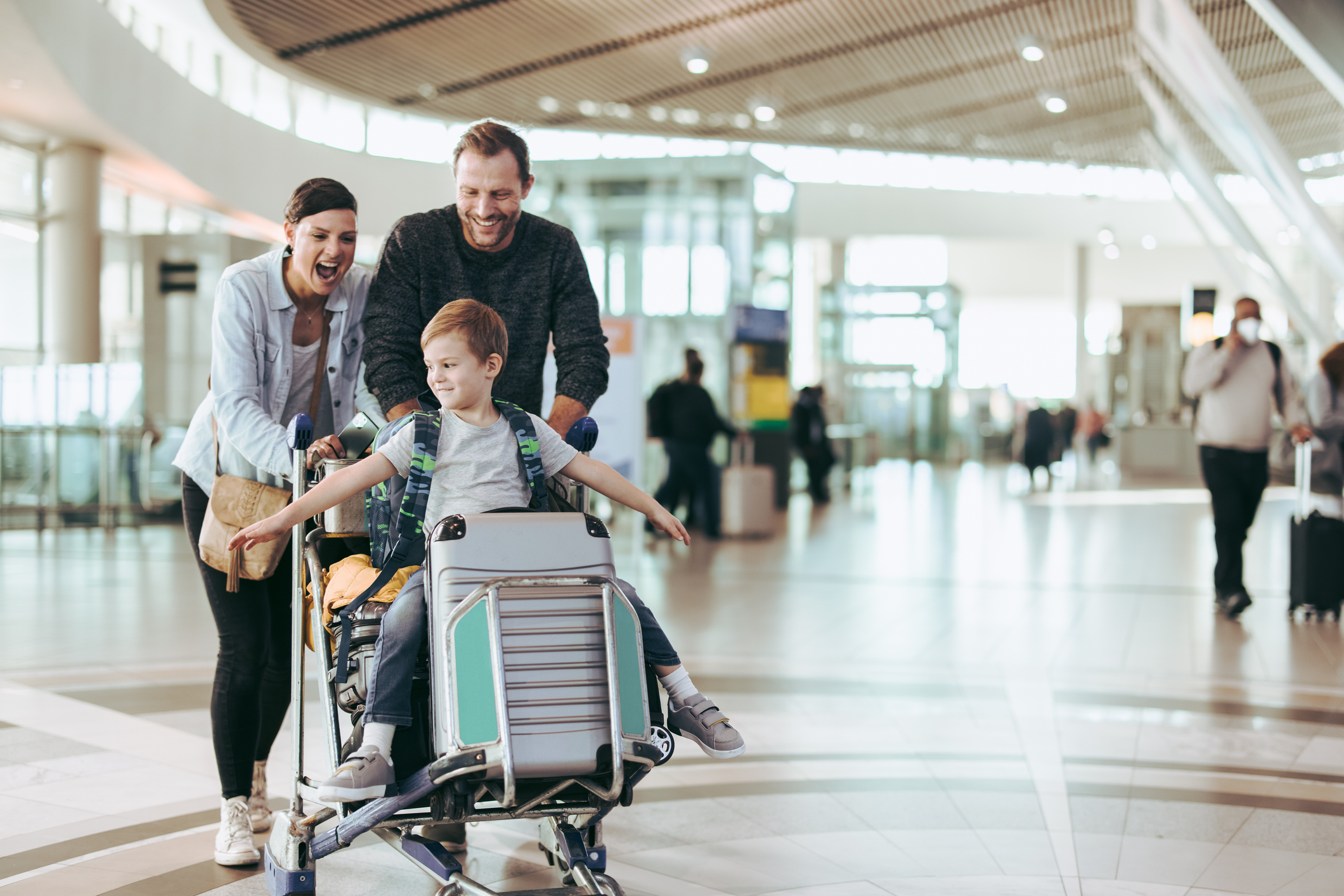 Familie schiebt Kind im Gepäckwagen durch Flughafen. Kind sitzt im Wagen und lächelt.